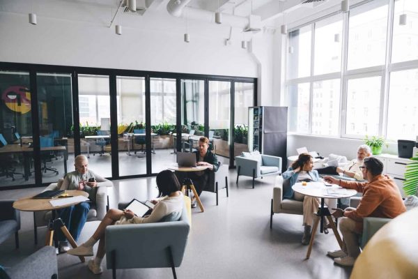 Full length of group of businesspeople with gadgets sitting in armchairs and talking while resting in cafeteria together during break in modern business center